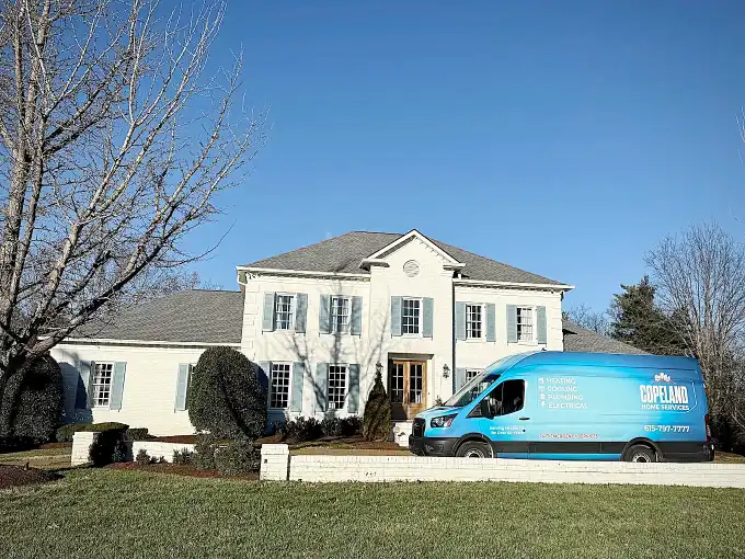A blue Copeland Home Services van is parked in front of a large two-story white house with columns on a sunny day.